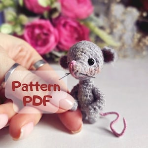 May include: A crocheted gray mouse amigurumi toy with pink ears and nose. The toy is being held by a hand with the text "Pattern PDF" on the finger.