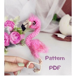 Puede incluir: Un flamenco rosa hecho a mano con un pico negro y una pata beige. El flamenco está hecho de hilo y tiene una textura esponjosa. El texto "Pattern PDF" es visible en la imagen.