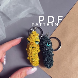 May include: Two handmade crochet keyrings, one yellow and one dark green, each with a whimsical creature design and attached to a silver keyring. The text "PDF PATTERN" is visible in the background.