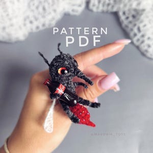 Puede incluir: Un insecto negro tejido a ganchillo con detalles rojos y blancos, sosteniendo una botella de Coca-Cola en miniatura. El insecto tiene grandes ojos naranjas y una cola roja. Incluye el texto "PATTERN PDF".