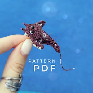 May include: A crocheted brown stingray with white spots and iridescent beads. The stingray is being held by a finger and has a long tail. The text "PATTERN PDF" is written below the stingray.