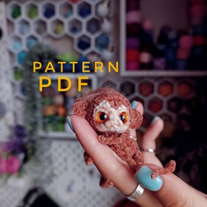 Könnte beinhalten: Ein gehäkelter brauner Affe mit großen orangefarbenen Augen wird in einer Hand mit blauem Nagellack gehalten. Der Affe hat ein hellbraunes Gesicht und ist mit "PATTERN PDF" in gelbem Text beschriftet.