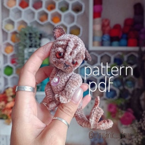 Puede incluir: Un juguete de peluche de crochet en forma de gato con un patrón de rayas marrones y blancas. El gato sostiene un pequeño amuleto de concha. El juguete está siendo sostenido por una mano y el texto "pattern pdf" es visible en el fondo.