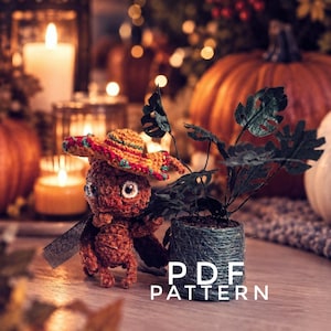 May include: A brown crocheted creature wearing a small orange sombrero with green and red trim, standing next to a miniature potted plant with dark green leaves. The text PDF PATTERN is printed in white letters on the lower right side.
