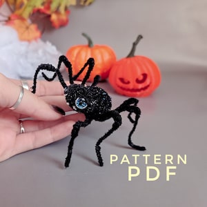 May include: Black spider craft pattern with sparkly black body and blue eyes. The spider is made with pipe cleaners and is sitting on a gray surface with two orange pumpkins in the background. The text "PATTERN PDF" is written in yellow on the gray surface.