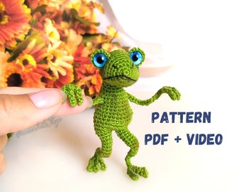 May include: A green crocheted frog with blue eyes. The frog is standing on a white surface. The text "PATTERN PDF + VIDEO" is in the lower right corner of the image.