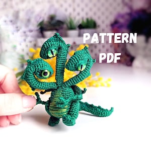 Può includere: Amigurumi di drago a tre teste di colore verde all'uncinetto con occhi verdi. L'amigurumi è in piedi su una superficie bianca. MODELLO PDF