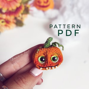 May include: A crocheted orange pumpkin with a green stem and googly eyes. The pumpkin has a toothy grin and is being held in a hand. The text "PATTERN PDF" is written in green letters on a white background.