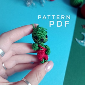 Puede incluir: Un juguete amigurumi de crochet verde con un mono rojo. El juguete tiene ojos negros y una nariz amarilla. El juguete está siendo sostenido en una mano. El texto "PATTERN PDF" es visible en el fondo.