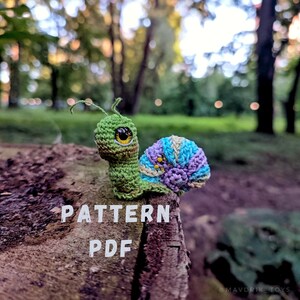 May include: Crochet pattern PDF for a green and purple snail amigurumi toy. The snail is sitting on a tree stump with the words "PATTERN PDF" written on the stump.