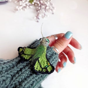 K&ouml;nnte beinhalten: Ein handgefertigter gr&uuml;ner und schwarzer H&auml;kel-Schmetterling mit Perlenakzenten. Der Schmetterling hat gro&szlig;e gr&uuml;ne Augen und ist an einem grauen Strick&auml;rmel befestigt. Das Bild ist aus der N&auml;he aufgenommen.