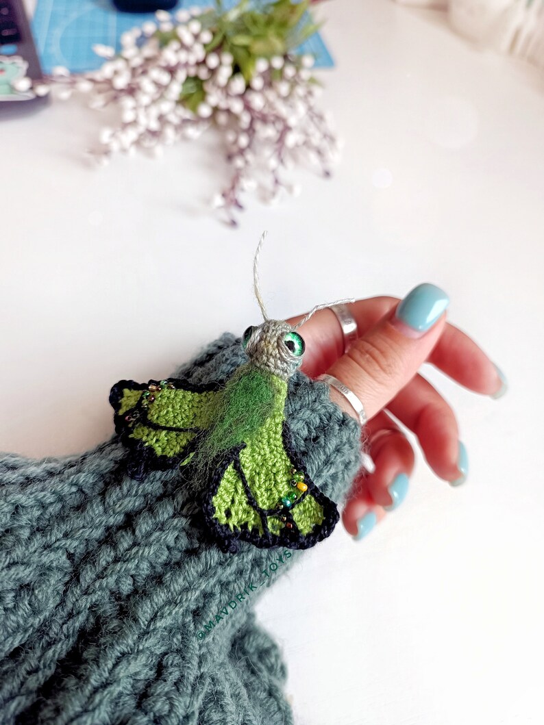K&ouml;nnte beinhalten: Ein handgefertigter gr&uuml;ner und schwarzer H&auml;kel-Schmetterling mit Perlenakzenten. Der Schmetterling hat gro&szlig;e gr&uuml;ne Augen und ist an einem grauen Strick&auml;rmel befestigt. Das Bild ist aus der N&auml;he aufgenommen.