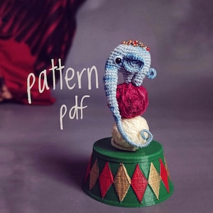 Puede incluir: Un juguete de elefante de crochet azul equilibrado sobre una pila de ovillos de lana en un pedestal de circo verde y rojo. El texto "pattern pdf" está escrito sobre un fondo borroso.
