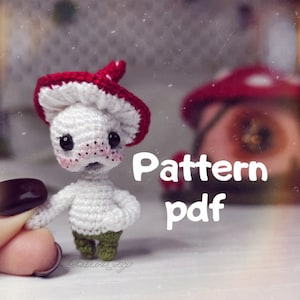 May include: A crocheted mushroom amigurumi toy with a red and white hat. The toy has a white body, green pants, and black eyes. The text "Pattern pdf" is written in white on a red background.