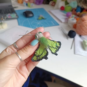 K&ouml;nnte beinhalten: Ein handgefertigter H&auml;kel-Schmetterling mit gr&uuml;nen Fl&uuml;geln, schwarzem Rand und Perlenakzenten. Der Schmetterling hat einen grauen Kopf und F&uuml;hler sowie gr&uuml;ne Augen. Die Fl&uuml;gel sind leuchtend gr&uuml;n mit einem schwarzen Rand und kleinen Perlendetails.