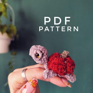 May include: A small, crocheted turtle figurine with a light purple head and limbs, featuring a bright red, textured shell that resembles an apple. The text "PDF PATTERN" is displayed in white letters above the handmade plush toy.