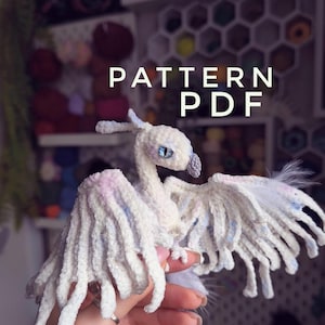 Puede incluir: Un pájaro blanco tejido a mano con alas extendidas, con detalles en azul y rosa. El pájaro tiene una cabeza detallada con ojos azules y un pico gris. La imagen incluye el texto "PATTERN PDF".