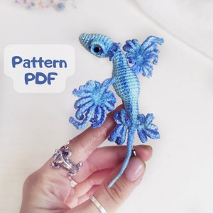 May include: A crocheted blue and light blue dragon with a long tail and large wings. The dragon is being held by a hand wearing a silver ring.