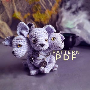Puede incluir: Un perro Cerbero de ganchillo de color lavanda con tres cabezas, cada una con ojos dorados. El peluche tiene un collar de cadena plateada con un colgante. La imagen incluye el texto "PATTERN PDF".