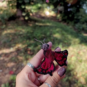 K&ouml;nnte beinhalten: Ein handgefertigter, miniaturisierter Schmetterling mit einem dunkelroten K&ouml;rper und schwarz umrandeten Fl&uuml;geln. Die Fl&uuml;gel sind mit kleinen, glitzernden Akzenten verziert. Der Schmetterling hat einen detaillierten Kopf und feine Antennen. Das Objekt wird in einer Hand vor einem verschwommenen gr&uuml;nen Hintergrund gehalten.