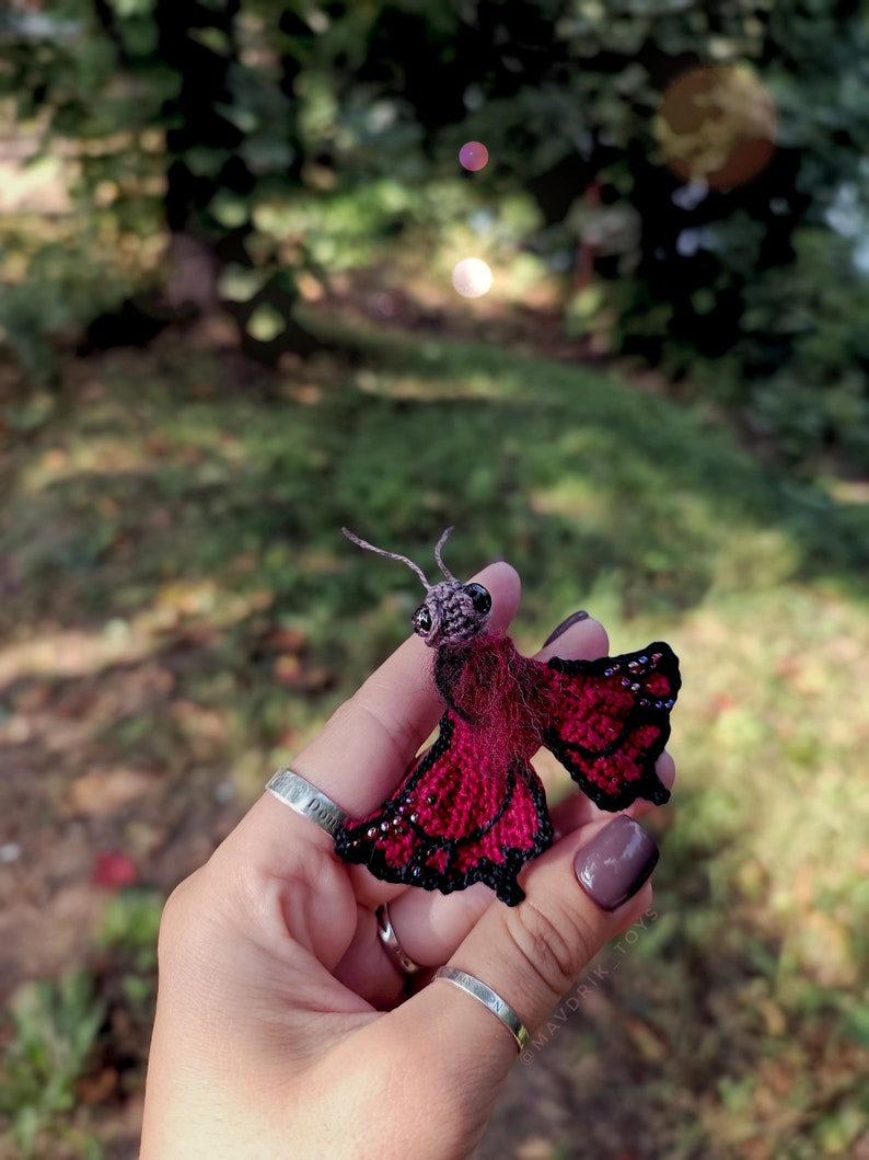 K&ouml;nnte beinhalten: Ein handgefertigter, miniaturisierter Schmetterling mit einem dunkelroten K&ouml;rper und schwarz umrandeten Fl&uuml;geln. Die Fl&uuml;gel sind mit kleinen, glitzernden Akzenten verziert. Der Schmetterling hat einen detaillierten Kopf und feine Antennen. Das Objekt wird in einer Hand vor einem verschwommenen gr&uuml;nen Hintergrund gehalten.