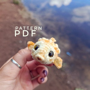 Könnte beinhalten: Ein gehäkeltes Amigurumi-Fischspielzeug mit orangefarbenem und weißem Fell, großen schwarzen Augen und einer kleinen rosa Nase. Das Spielzeug wird von einer Hand mit einem silbernen Ring gehalten. Der Hintergrund ist ein verschwommenes Bild eines Berges und des Himmels.