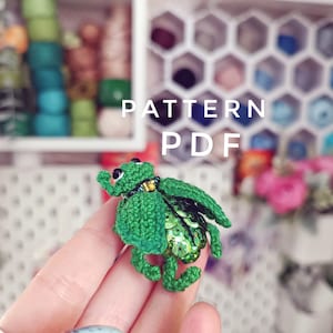 Puede incluir: Un escarabajo verde tejido a crochet con lentejuelas verdes en la espalda. El escarabajo está siendo sostenido por una mano. El texto "PATTERN PDF" es visible en el fondo.