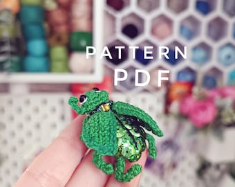 May include: A green crocheted beetle with green sequins on its back. The beetle is being held by a hand. The text "PATTERN PDF" is visible in the background.