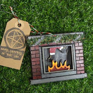 May include: A wooden Christmas ornament shaped like a fireplace with a Santa Claus leg sticking out of the chimney. The fireplace is painted red, black, and gray with a yellow flame inside. The ornament is decorated with a green garland.