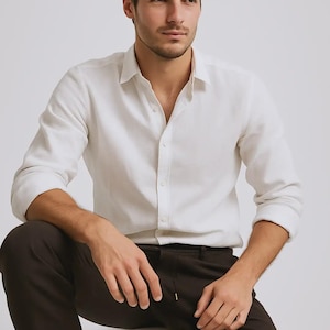 Pode incluir: Uma camisa branca de manga comprida e calças castanhas escuras. A camisa é feita de tecido leve e tem uma gola clássica. As calças são de cor lisa e têm um cordão. O conjunto é casual e elegante.