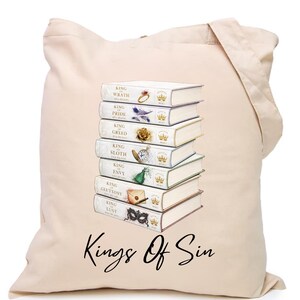 May include: A beige canvas tote bag featuring a stack of seven white books with gold crown icons and the text Kings Of Sin printed below. Each book spine displays a title and a unique symbol, including a ring, feather, rose, watch, gem, envelope, and mask.
