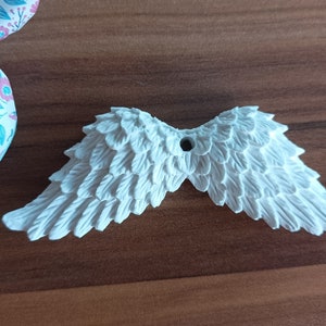 Peut inclure: Ornement en forme d'ailes d'ange blanches et détaillées, avec un trou pour l'accrocher. L'aile est faite d'un matériau texturé et est posée sur une surface en bois. Un ruban à motifs floraux est partiellement visible en haut à gauche.