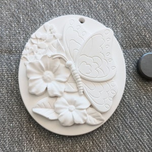 May include: A white oval-shaped plaque featuring a raised relief design of a butterfly resting on flowers and leaves. The plaque has a small hole at the top for hanging and is accompanied by a separate small, round, dark gray magnet.