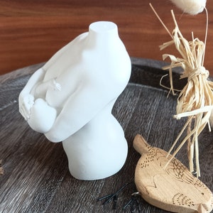 Könnte beinhalten: Weiße Gipsfigur einer Frau, die ein Baby hält. Die Skulptur steht auf einer Holzoberfläche mit einem kleinen Holzvogel und einigen schwarzen Haarnadeln.