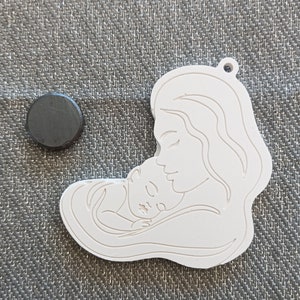 以下が含まれることがあります： 母親が眠っている赤ちゃんを抱いている白いペンダント。上部に小さな穴が開いています。濃い灰色の円形磁石がペンダントの隣にあります。ペンダントはおそらく記念品または装飾品です。