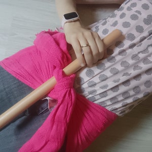 Op de afbeelding: Een persoon draagt ​​een roze sjaal die om de taille is geknoopt met een houten stok die op de sjaal ligt. De persoon draagt ​​een wit shirt met een bloemenpatroon en een blauwe jeans.
