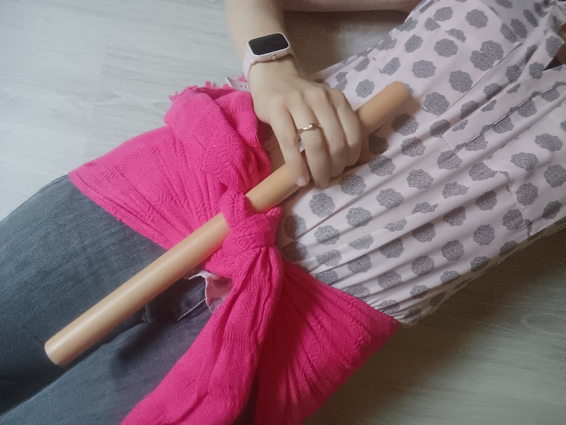 Op de afbeelding: Een persoon draagt ​​een roze sjaal die om de taille is geknoopt met een houten stok die op de sjaal ligt. De persoon draagt ​​een wit shirt met een bloemenpatroon en een blauwe jeans.