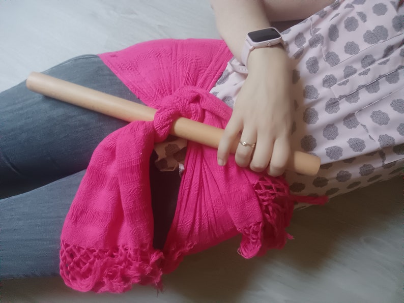 Op de afbeelding: Een roze geweven wikkel met een houten stok die erdoorheen is geregen. De wikkel is om de taille van een persoon geknoopt.