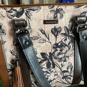 以下が含まれることがあります： 花柄のプリントと前面に「handmade」の文字が書かれた、茶色と黒のトートバッグ。バッグには、茶色のアクセントが付いた黒いストラップが2本と、茶色のタッセルが付いています。
