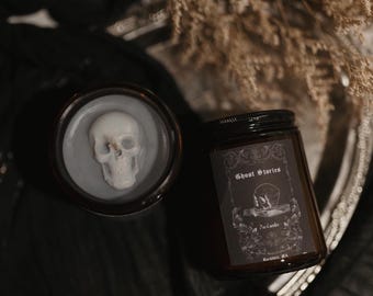May include: Two dark glass jars with black lids. One jar contains a white skull embedded in a gray wax. The other jar has a label that reads "Ghost Stories" with a skull and book illustration. The jars are on a silver tray.