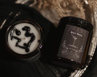 May include: Two glass jar candles with black lids. One candle has a black design on the white wax. The other candle has a label that reads "Black Honey" with a cocktail glass illustration. The candles are on a silver tray.
