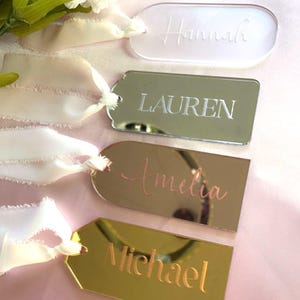 Puede incluir: Cuatro etiquetas de regalo de acrílico personalizadas con nombres grabados. Las etiquetas son de diferentes colores: transparente, plateado, oro rosa y oro. Las etiquetas están atadas con una cinta blanca.