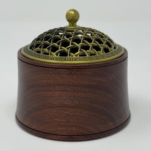 May include: A dark brown wooden incense burner with a decorative brass lid. The lid features a lattice design and a small brass knob. The burner has a round, cylindrical shape and is likely used for aromatherapy or meditation.