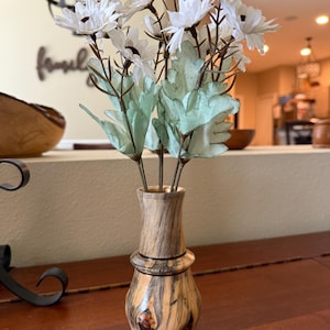 May include: A wooden vase with a light brown and dark brown grain pattern holds white daisy flowers with brown centers and green leaves. The vase has a bulbous base and a narrow neck, and sits on a dark wooden surface.