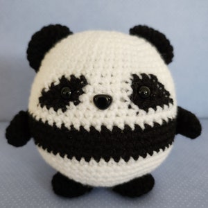 Pode incluir: Um bicho de pelúcia de urso panda feito à mão em crochê. O panda tem um corpo branco com detalhes pretos ao redor dos olhos, orelhas e membros. Possui olhos de botão pretos e um pequeno nariz preto. O brinquedo é redondo e tem uma aparência fofa e amigável.