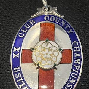 Puede incluir: Medalla de plata con esmalte azul y el texto "English Club County Championship XX" alrededor del borde. Una cruz roja con una rosa blanca en el centro está en la medalla.