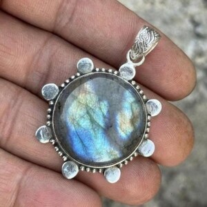 May include: A round pendant featuring a polished labradorite gemstone with iridescent blue and gold flashes. The stone is set in a silver-toned metal frame decorated with small circular accents along the edge and a textured, patterned bail at the top.