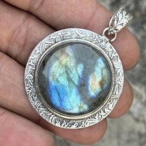 May include: A round pendant featuring a polished, iridescent labradorite gemstone with blue and gold flashes, set within a silver-toned metal frame decorated with intricate, embossed floral and vine patterns and a matching ornate bail.