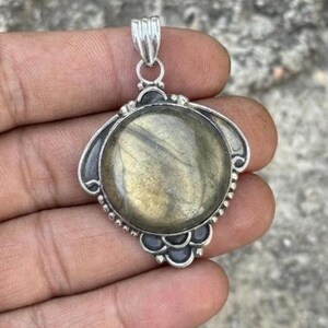 May include: A round labradorite gemstone pendant set in a silver frame with decorative metal accents. The stone displays a muted golden-green iridescence within a polished circular setting, featuring a silver bail at the top for hanging.
