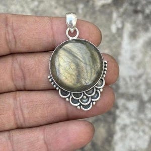 May include: A round labradorite gemstone pendant set in a silver frame. The stone displays a muted grey and gold iridescence. The silver setting features a decorative scalloped edge along the bottom half and a simple bail at the top.